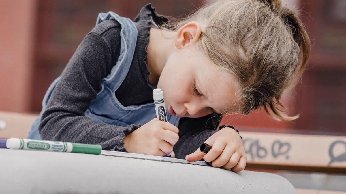 Adorable concentrated little girl in denim overall drawing picture with color markers on paper