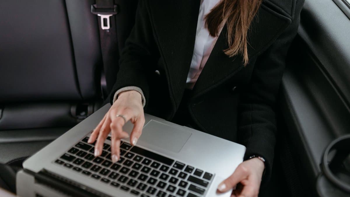 Businesswoman in black blazer using laptop inside a car, focused on work.