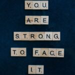 Scrabble tiles spelling 'You Are Strong To Face It' on a dark surface, conveying empowerment.
