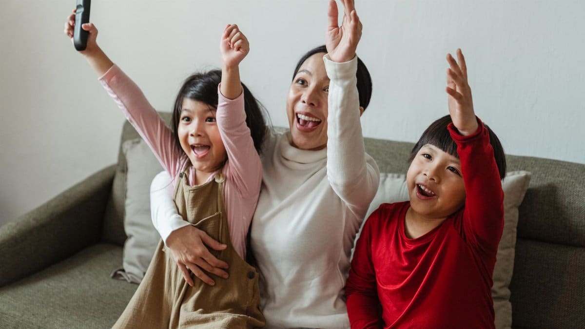Cheerful Asian mom and kids watching sport on TV at home and screaming happily while cheering with raised arms