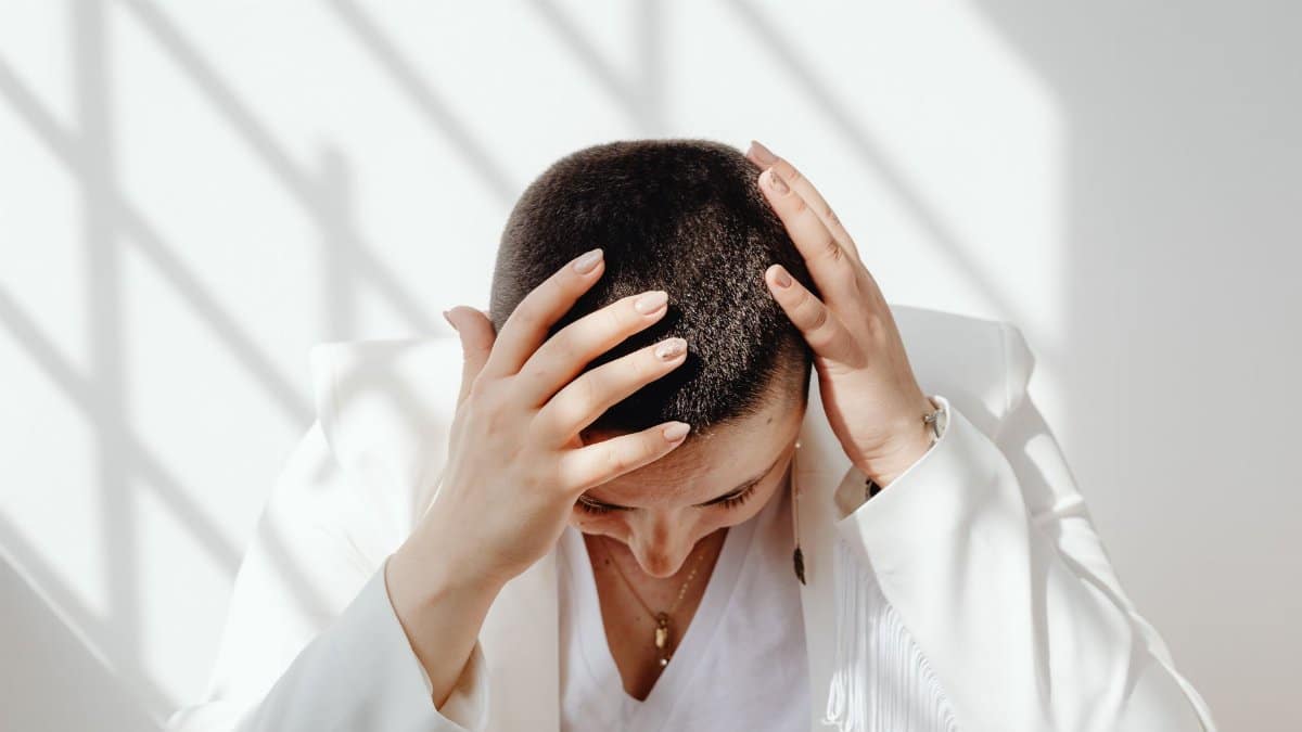 Woman in white outfit feeling stressed, sitting with hands on head in bright sunlight indoors.
