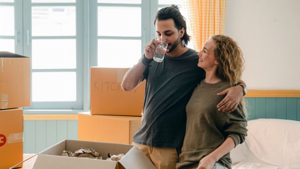 Ethnic man drinking water while cuddling happy girlfriend standing near pile of cardboard boxes in sunlight after moving into new apartment