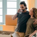 Ethnic man drinking water while cuddling happy girlfriend standing near pile of cardboard boxes in sunlight after moving into new apartment
