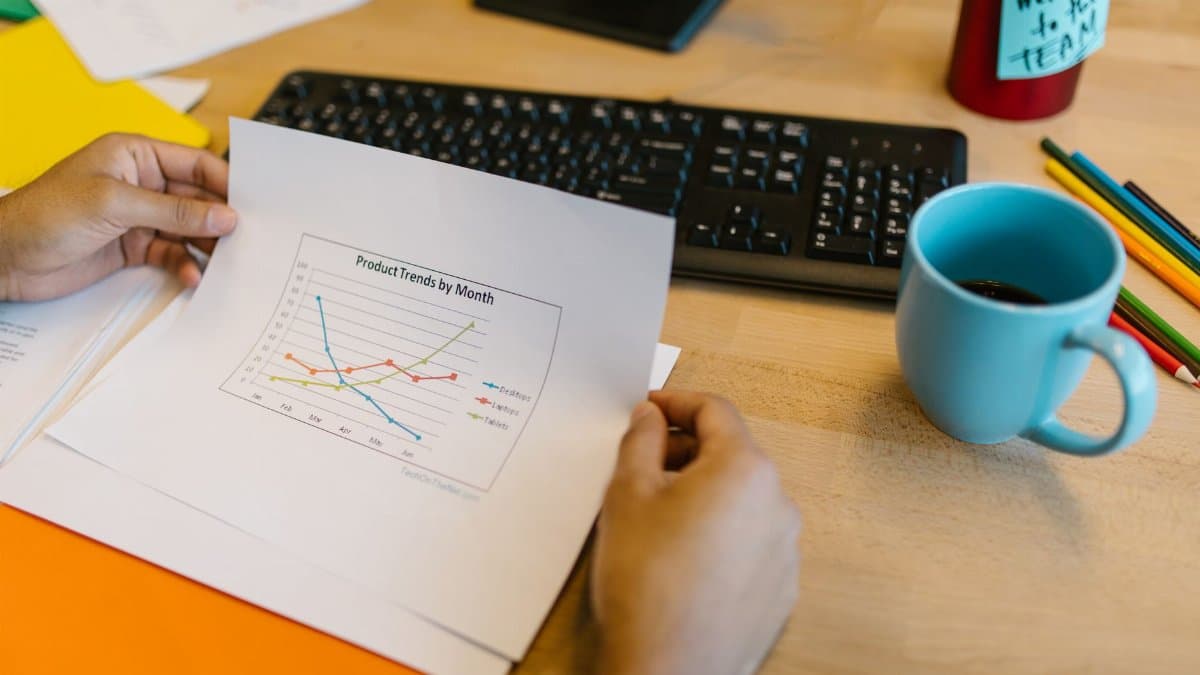 Close-up of hands reviewing a paper graph with a coffee cup and keyboard on a desk.