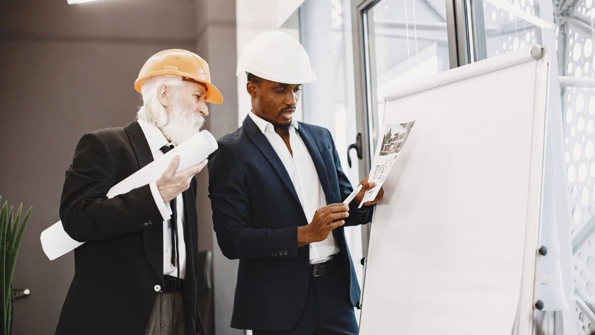 Two architects in suits and hard hats discussing architectural plans on a flipchart.