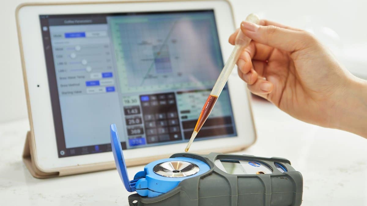 Hand using pipette with refractometer in front of tablet displaying measurement interface.