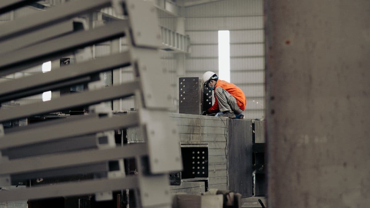 Worker in orange safety suit and white helmet inspecting metal structures in a factory.