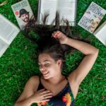 A young woman in a floral dress smiling while lying on grass with open books around her.