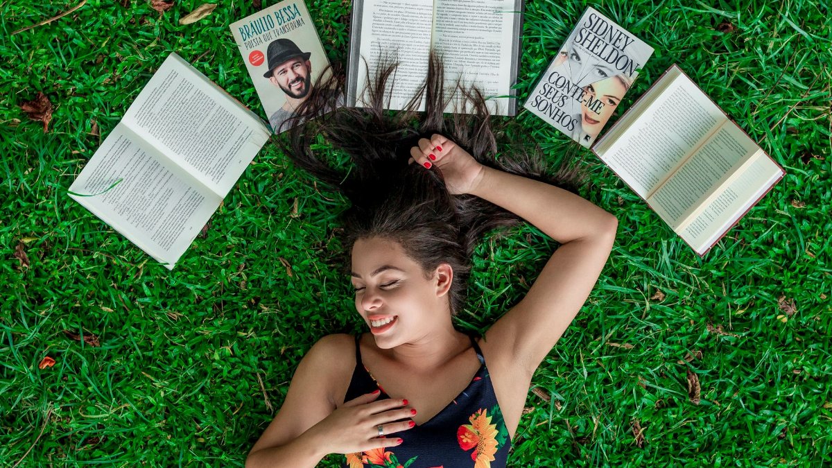 A young woman in a floral dress smiling while lying on grass with open books around her.