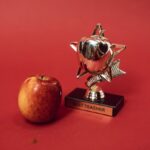 A 'Best Teacher' trophy alongside an apple on a vivid red background symbolizing education and recognition.