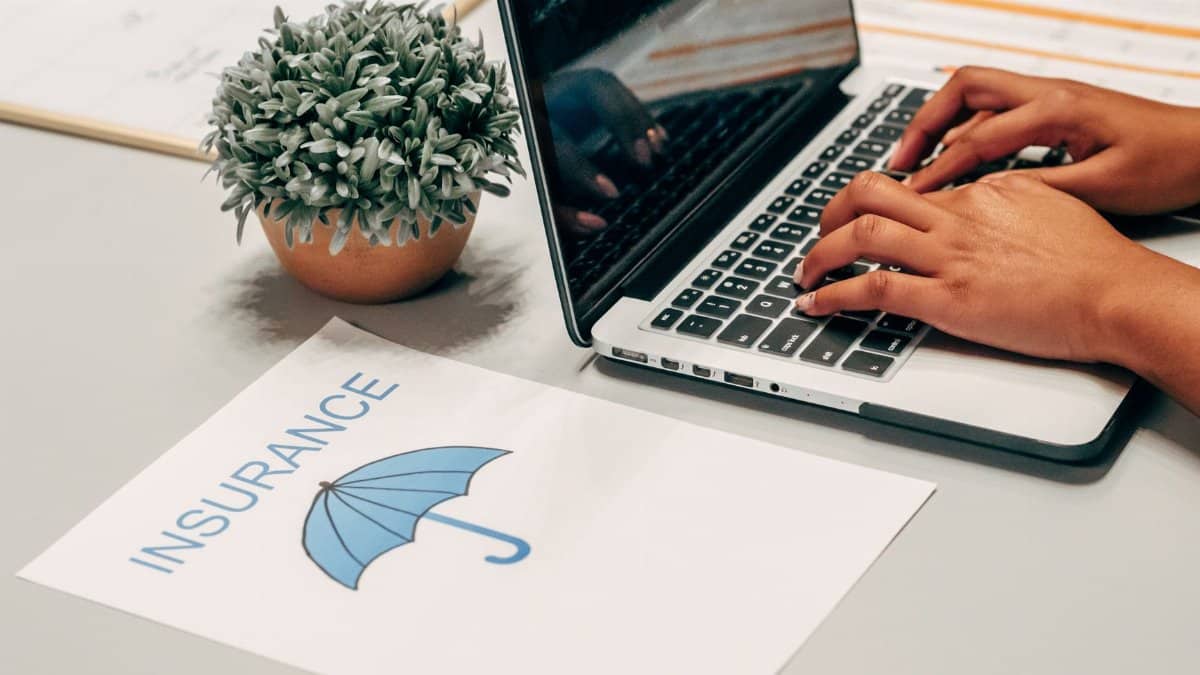 Hands typing on a laptop at a desk with an insurance paper and plant, suggesting a work environment.