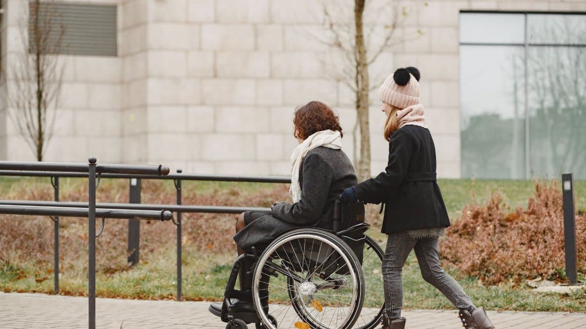 A young girl helps a woman in a wheelchair on a sidewalk, showing care and support.