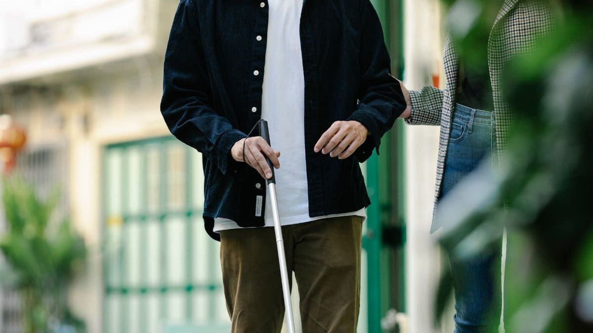 A young adult with visual impairment using a walking stick, aided by a companion outdoors.