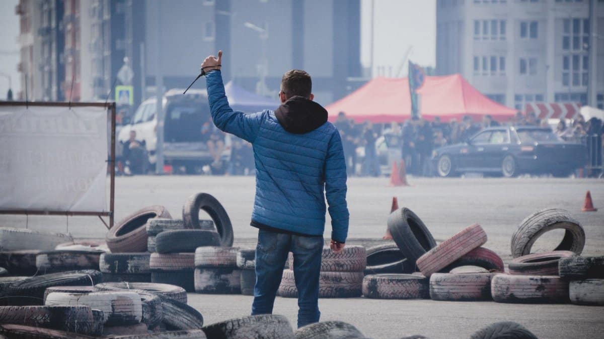 Man with blue jacket signals an urban car racing event amidst tire barriers and cityscapes.
