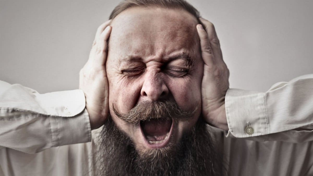 Portrait of a man with facial hair, covering his ears and screaming, expressing stress or frustration.