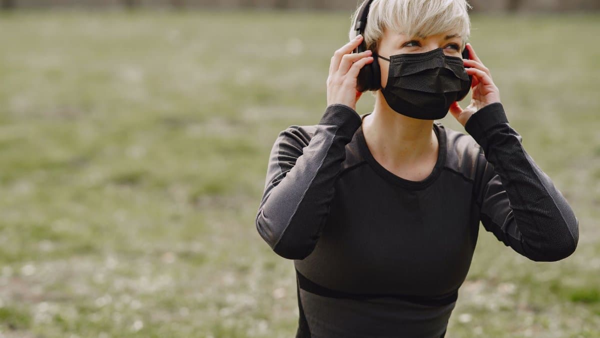 Young female in sportswear with black protective mask looking away and adjusting headphones while having rest after working out in park