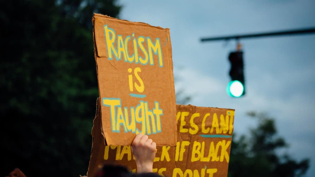 Crop anonymous activist demonstrating placard with racism is taught text while protesting for Black Lives Matter movement in evening
