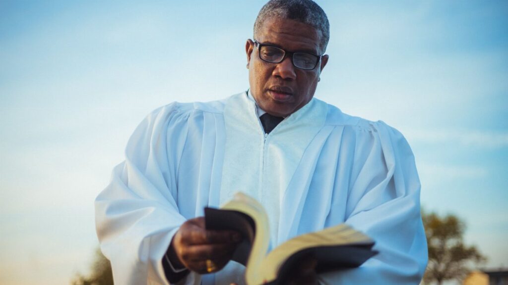 An adult pastor wearing glasses and a white robe reads a book outdoors under a blue sky.