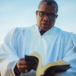 An adult pastor wearing glasses and a white robe reads a book outdoors under a blue sky.