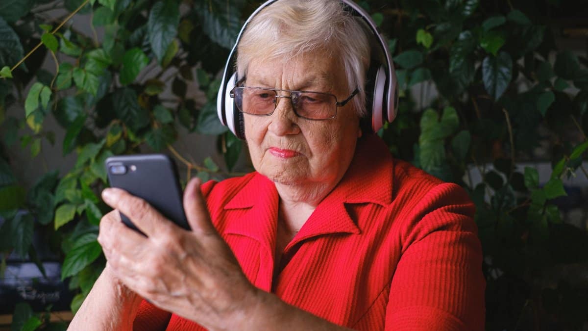 Concentrated senior female wearing eyeglasses and headphones listening to music while surfing cellphone in room near green leaves during free time