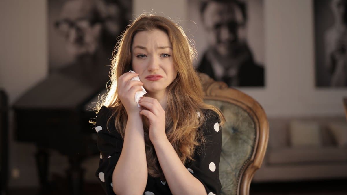 A woman sitting in a vintage chair indoors, showing emotions and holding tissues.