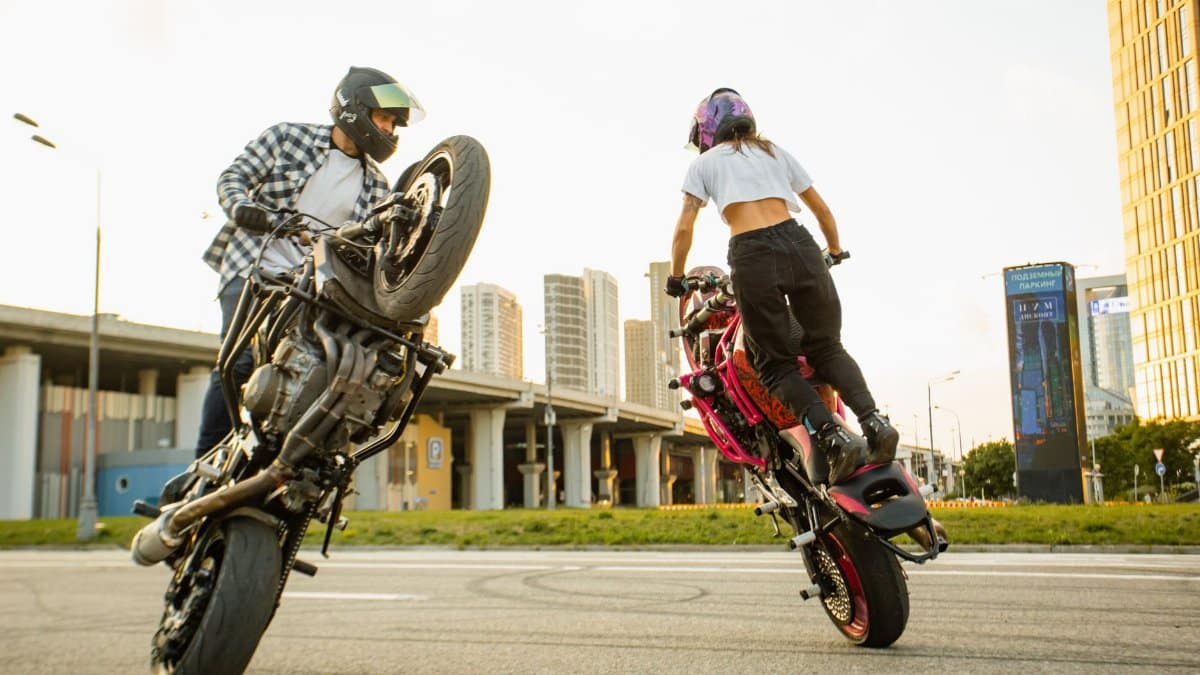 Two riders performing motorcycle stunts in a cityscape setting.