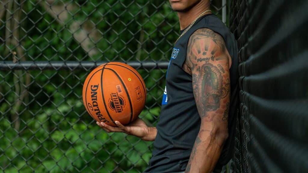 Athletic adult man holding basketball against a fence outdoors, showcasing tattoos and sports attire.