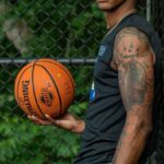 Athletic adult man holding basketball against a fence outdoors, showcasing tattoos and sports attire.