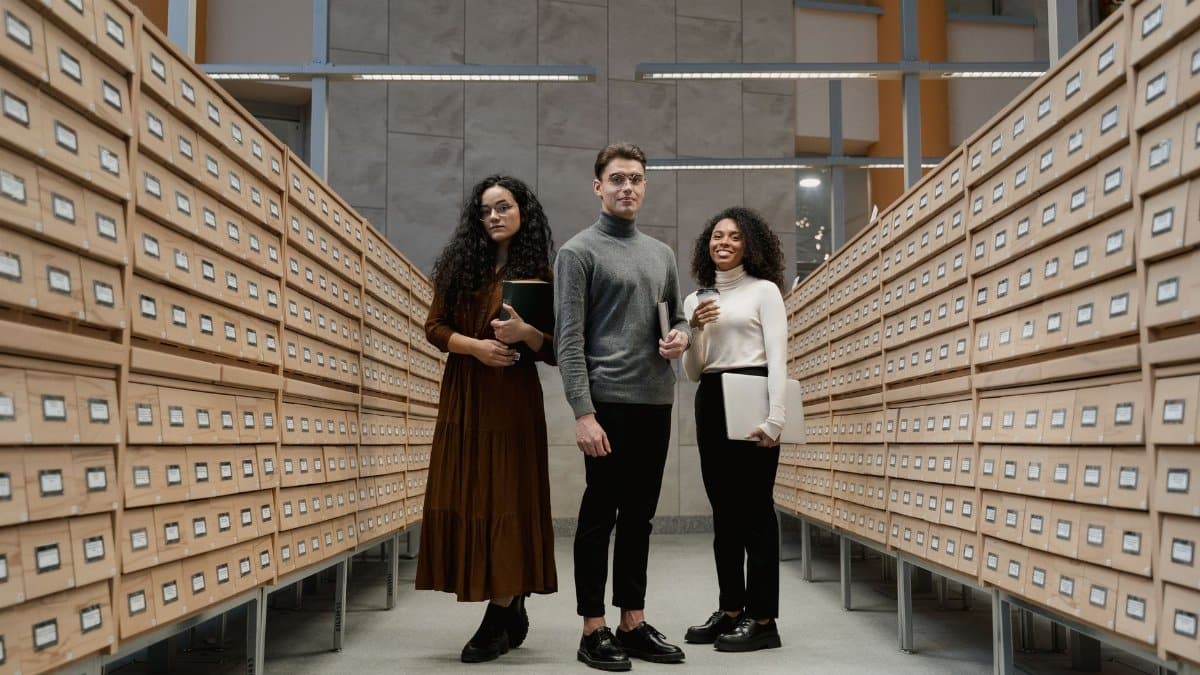 Three students stand in a library archive, symbolizing diversity and learning.