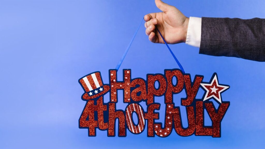 Hand holding glittery 4th of July sign with stars and stripes on blue backdrop.