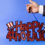 Hand holding glittery 4th of July sign with stars and stripes on blue backdrop.