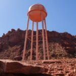 A solitary water tower stands against a rugged rocky desert backdrop under a clear blue sky.