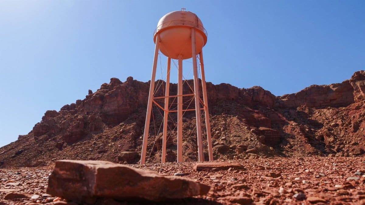 A solitary water tower stands against a rugged rocky desert backdrop under a clear blue sky.