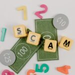 Scrabble tiles spelling 'scam' with play money and numbers on a white background.