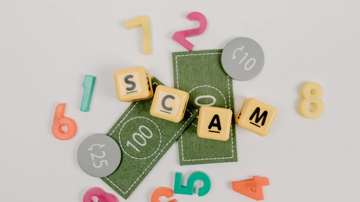 Scrabble tiles spelling 'scam' with play money and numbers on a white background.
