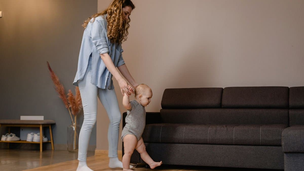 A mother supports her baby as they take first steps near a sofa indoors, fostering childhood development.