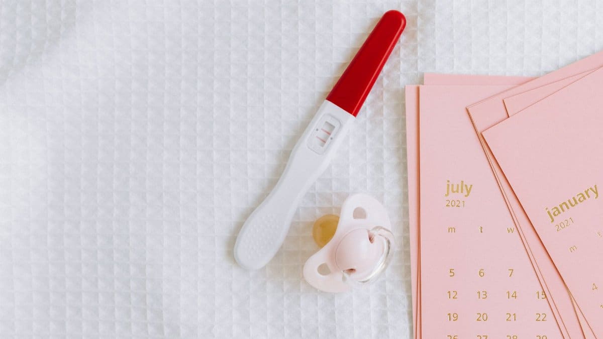 A pregnancy test with a positive result next to a baby pacifier and calendar on a textured surface.