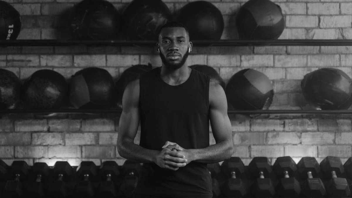 Monochrome image of a fit man in a gym surrounded by medicine balls and dumbbells.