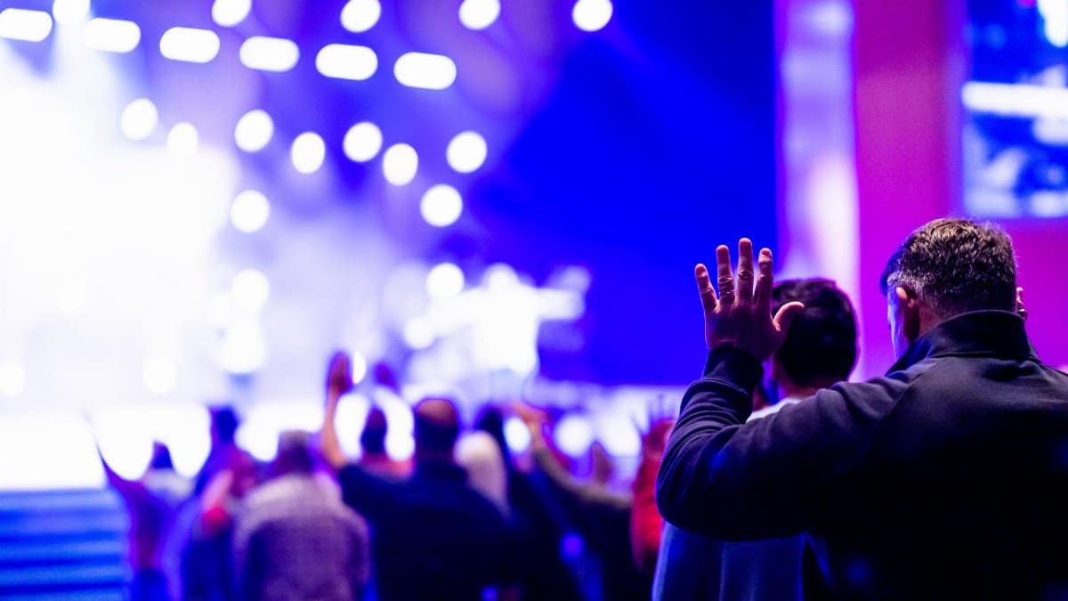 A lively and engaging indoor worship celebration filled with a diverse crowd immersed in prayer and music under bright stage lights.