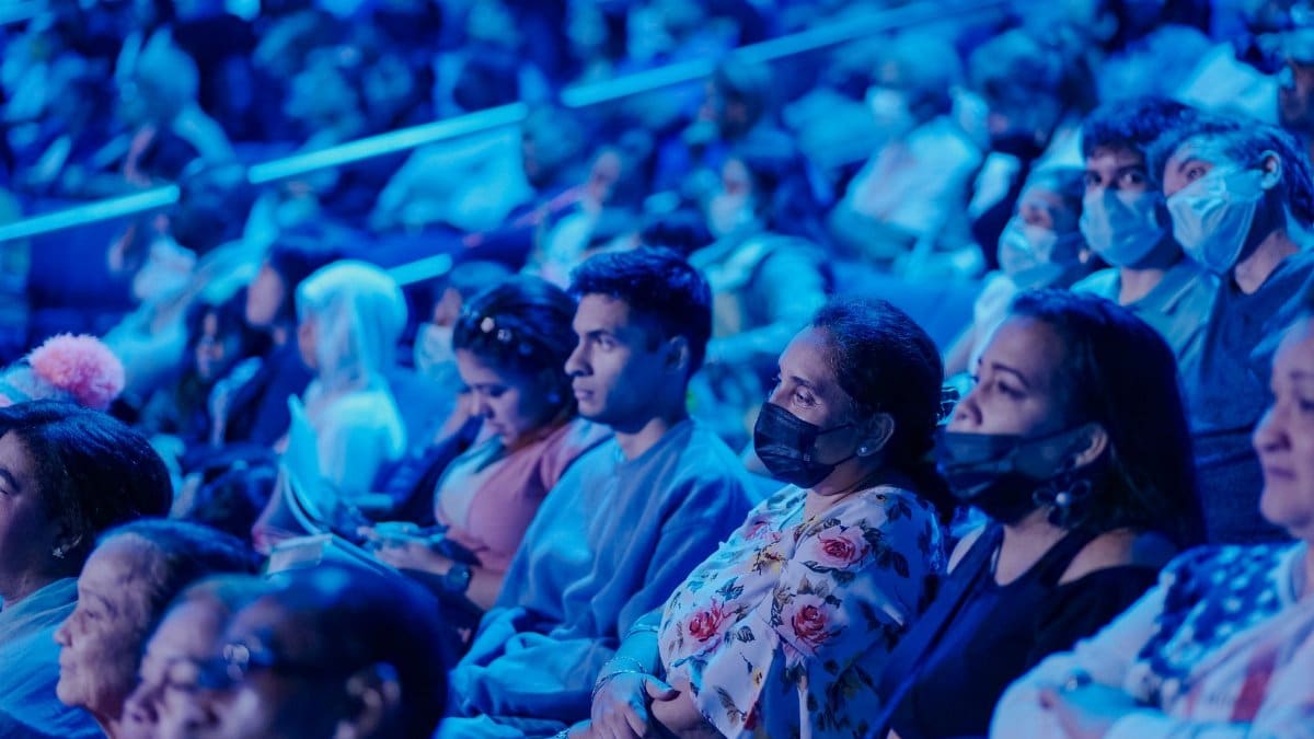 A diverse audience in masks attentively watching a concert under blue lights.