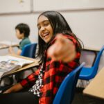Young student in a plaid shirt smiling and pointing playfully in a classroom setting.