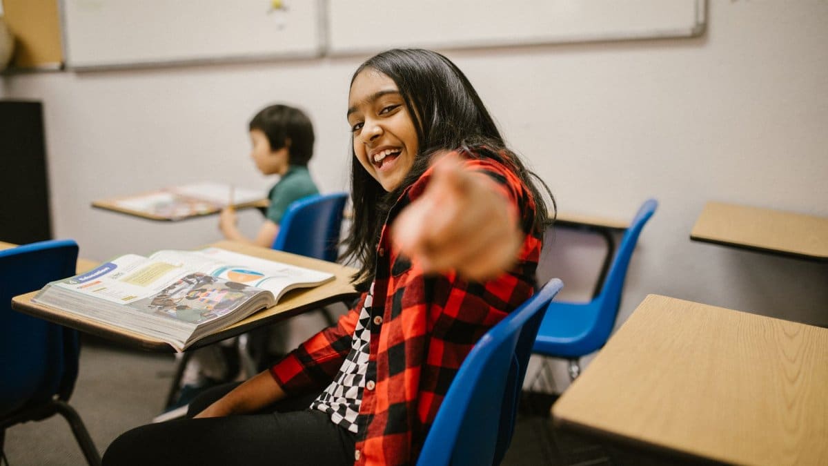 Young student in a plaid shirt smiling and pointing playfully in a classroom setting.