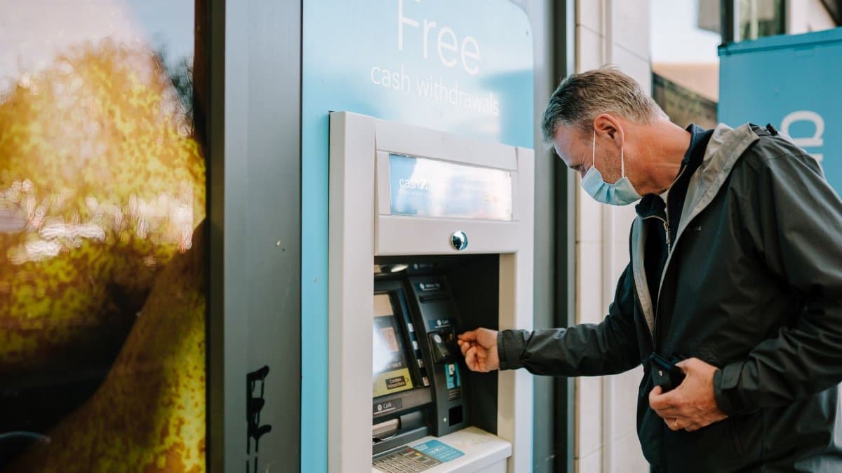 A man wearing a mask using an ATM machine outdoors for cash withdrawal.