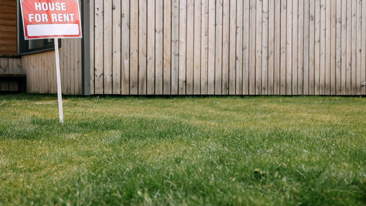 Close-up of 'House for Rent' sign on green lawn in front of a wooden house exterior.