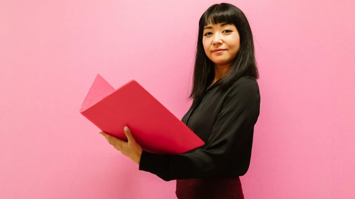 Smiling woman in business attire holding a pink folder against a pink background.