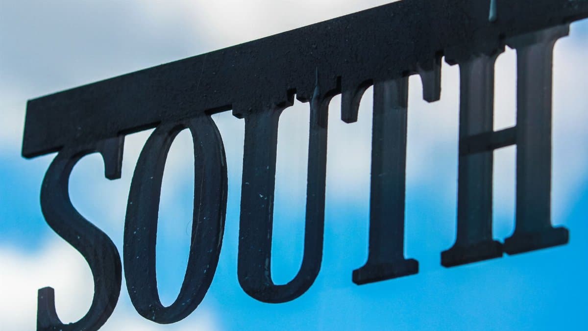 Minimalist metal 'south' sign silhouetted against a bright blue sky with cloud background.