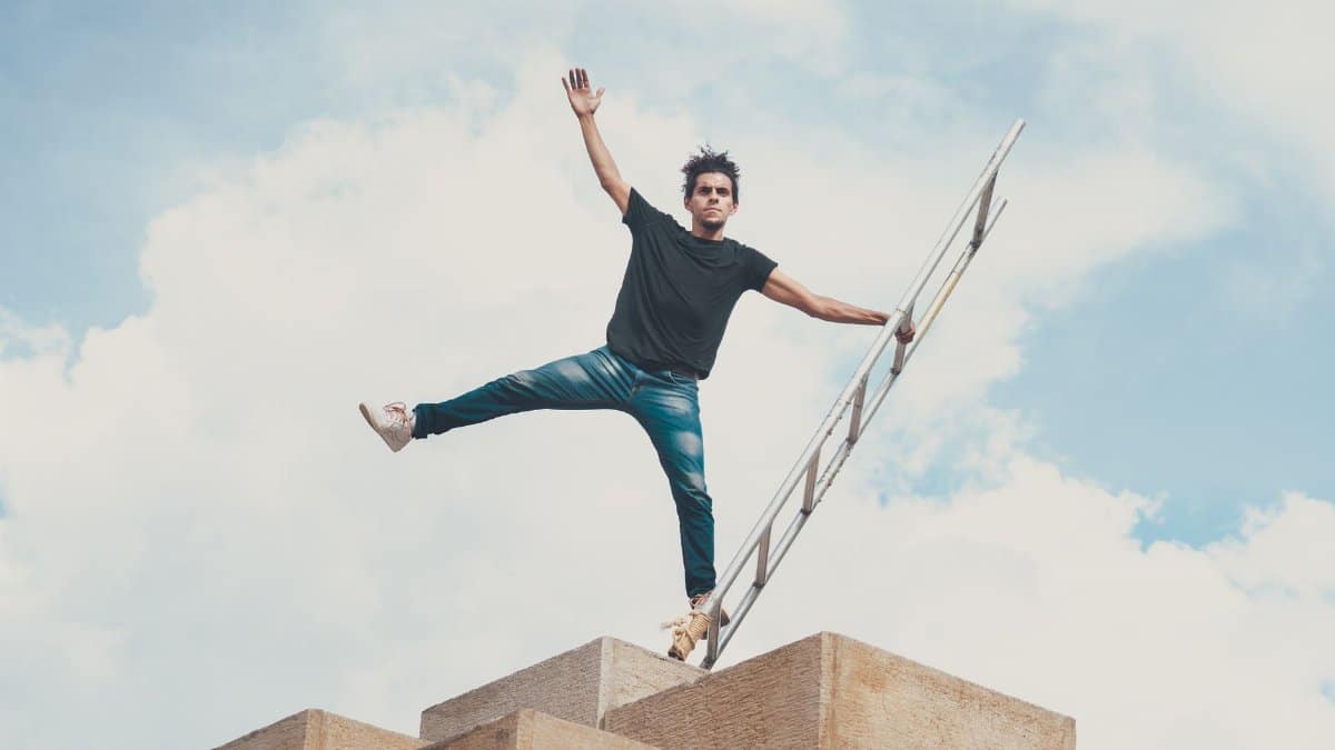 Man balancing on a ladder up high against a vibrant blue sky with clouds.