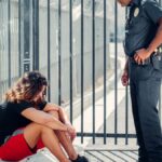A police officer stands over a seated civilian by a metal fence, illustrating authority and interaction.