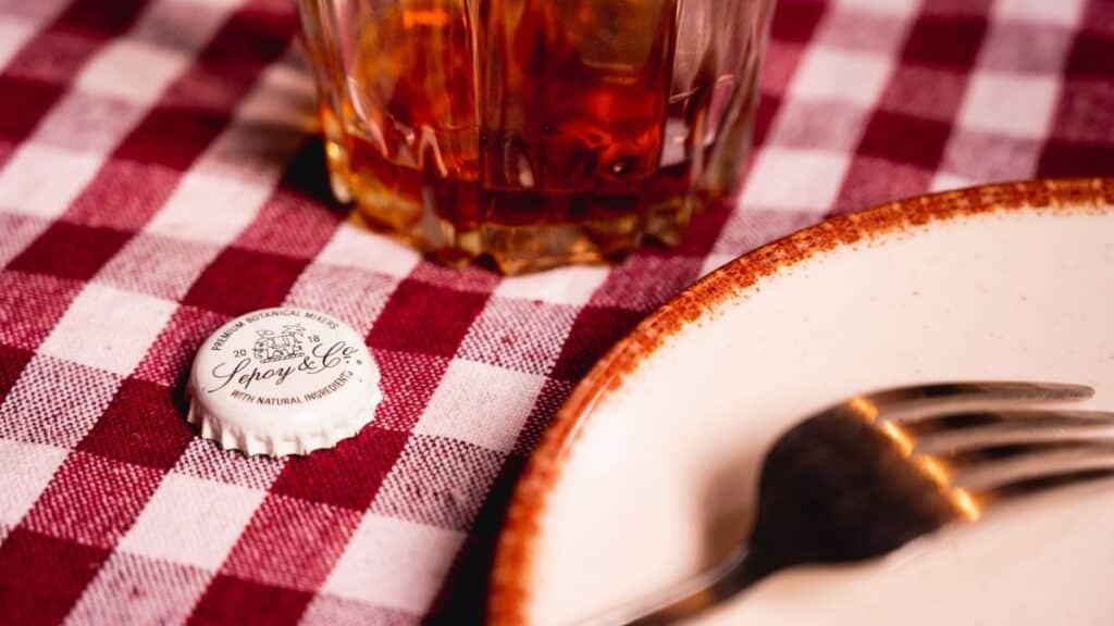 A drink and bottle cap on a red checkered tablecloth with a fork and plate visible, creating a rustic dining ambiance.