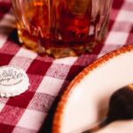 A drink and bottle cap on a red checkered tablecloth with a fork and plate visible, creating a rustic dining ambiance.